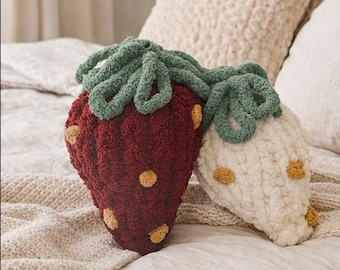 Op de afbeelding: Twee aardbei-vormige sierkussens. Eén is dieprood met gele zaaddetails en groene bladeren, de andere is gebroken wit met gele zaden en groene bladeren. De kussens liggen op een bed met neutraal gekleurd beddengoed.