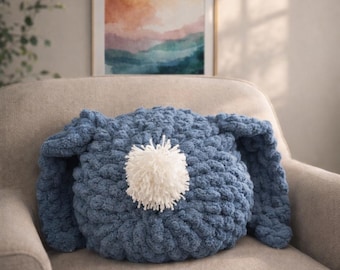 Op de afbeelding: Een blauw, konijnvormig kussen met lange oren en een witte pompon in het midden. Het kussen is gemaakt van een dik gebreid materiaal en ligt op een beige fauteuil. Het ontwerp van het kussen is grillig en decoratief.