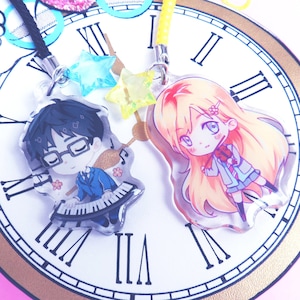 Può includere: Due ciondoli di personaggi anime, uno di un ragazzo con capelli neri e occhiali che suona una tastiera, e l'altro di una ragazza con lunghi capelli biondi, entrambi in stile chibi. I ciondoli sono appesi a un quadrante dell'orologio.