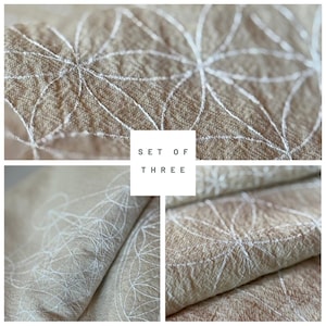 May include: A set of three beige fabric pieces with white embroidered floral patterns. The fabric has a textured look, and the text "SET OF THREE" is displayed in a white square.