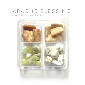 May include: A clear plastic container with four compartments, each filled with different coloured tumbled stones. The compartments contain light brown, light pink, light green, and white stones. The container is labelled "APACHE BLESSING CRYSTAL COLLECTION".