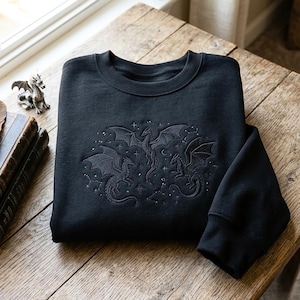 Könnte beinhalten: Ein schwarzes Sweatshirt mit Rundhalsausschnitt und einer dunklen Ton-in-Ton-Stickerei auf der Brust. Das Design zeigt drei geflügelte Drachen, umgeben von kleinen Sternen und Himmelsmustern. Der Stoff hat einen gerippten Kragen und Bündchen.