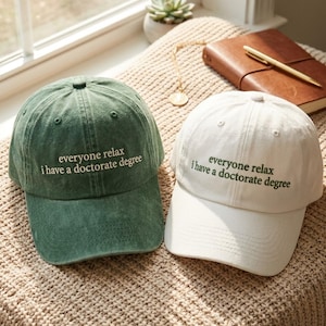 Puede incluir: Dos gorras de béisbol, una en verde lavado y otra en blanco, ambas con el texto bordado 'everyone relax i have a doctorate degree' en un color contrastante. Las gorras tienen una corona estructurada, una visera curva y ojales.