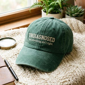Può includere: Un cappellino da baseball verde bosco con il testo ricamato in bianco "UNDIAGNOSED but something ain't right." Il cappellino presenta una corona strutturata, una visiera curva e occhielli cuciti sui pannelli superiori, dal design classico.