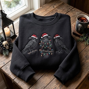 Peut inclure: Un sweat-shirt noir à col rond orné d'une broderie représentant trois corbeaux noirs. Chaque corbeau porte un bonnet de Père Noël rouge et blanc. Le corbeau central est perché sur une branche entourée de guirlandes lumineuses colorées.