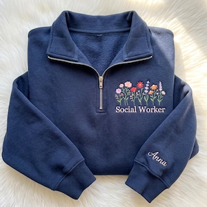 Könnte beinhalten: Ein marineblaues Sweatshirt mit kurzem Reißverschluss und einer Stickerei aus bunten Wildblumen über dem Text Social Worker in Hellrosa. Am rechten Ärmelbündchen ist der Name Anna in einer passenden hellrosa Schreibschrift eingestickt.