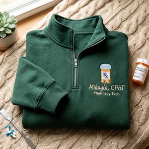 May include: A dark green quarter-zip pullover featuring an embroidered prescription bottle icon with the letters Rx, the name Mikayla, CPhT, and the text Pharmacy Tech. The garment has a silver zipper, long sleeves with ribbed cuffs, and a high collar.