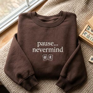 May include: A dark brown crewneck sweatshirt featuring the white embroidered text "pause... nevermind" centered on the chest, positioned above two small white rectangular icons depicting Mahjong tiles with Chinese characters.