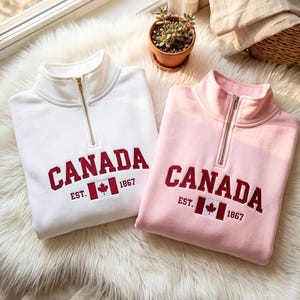 May include: A white and a light pink quarter-zip sweatshirt, each featuring the embroidered text CANADA EST. 1867 in dark red, accompanied by a small embroidered Canadian flag graphic centred below the text on the front of the garments.
