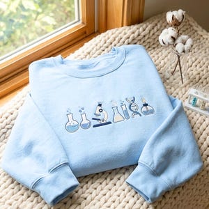 Peut inclure: Un sweat-shirt bleu clair à col rond avec une broderie sur la poitrine. Le motif représente divers équipements de laboratoire scientifique, notamment des béchers, un microscope, un tube à essai, une hélice d'ADN et une lampe à alcool.