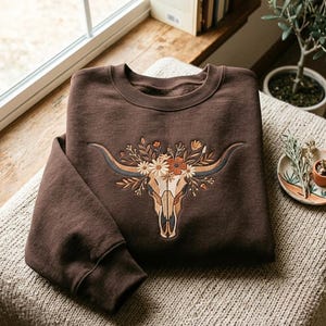 Op de afbeelding: Een donkerbruine sweater met ronde hals en een centrale geborduurde afbeelding van een longhorn-schedel, versierd met diverse bloemen en bladeren in tinten oranje, crème en beige. Het kledingstuk heeft lange mouwen en een geribbelde kraag en boorden.