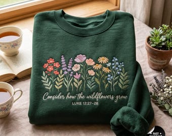 May include: A dark green crewneck sweatshirt featuring an embroidered design of colorful wildflowers in shades of pink, orange, yellow, and blue. Below the flowers, the text reads: Consider how the wildflowers grow, LUKE 12:27-28.