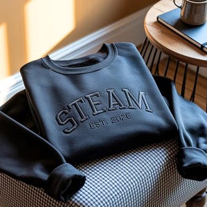 May include: A black long-sleeve crewneck sweatshirt featuring the word STEAM in large, arched, embroidered capital letters across the chest, with the text EST. 2026 embroidered in a smaller font directly underneath the main word.