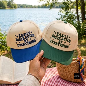 May include: Two baseball caps with cream-colored crowns and blue or green brims. Both caps feature the text "EASILY DISTRACTED BY EVERYTHING" in a dark blue or dark green font, accented with small four-pointed star symbols on the front panels.