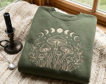 May include: An olive green crewneck sweatshirt featuring a light-colored line art graphic of various mushrooms, swirling vines, and a lunar cycle phase arc across the top. The design is centered on the front of the garment.