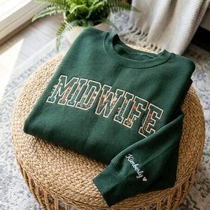 May include: A dark green crewneck sweatshirt featuring the word MIDWIFE embroidered in a bold, block-letter font with a colourful floral pattern inside the letters. The sleeve is embroidered with the name Kimberly and a small heart symbol.