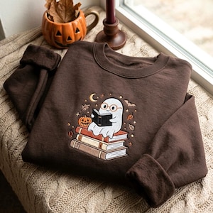 Peut inclure: Un sweat-shirt à col rond marron foncé avec une broderie représentant un fantôme blanc portant des lunettes rondes et lisant un livre noir, assis sur une pile de trois livres, accompagné d'une petite citrouille orange, de feuilles et d'une chauve-souris.
