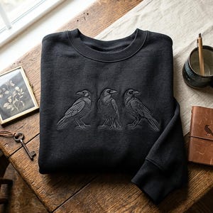 May include: A black crewneck sweatshirt featuring an embroidered design of three crows standing in a row. The birds are depicted in a detailed, monochromatic style with visible feather textures and sharp beaks, centered on the chest of the garment.