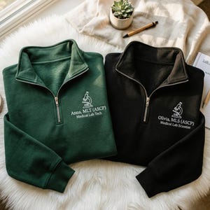 May include: A dark green and a black quarter-zip sweatshirt, each featuring a white embroidered microscope icon and text. The green sweatshirt reads Anna, MLT (ASCP) Medical Lab Tech, and the black sweatshirt reads Olivia, MLS (ASCP) Medical Lab Scientist.