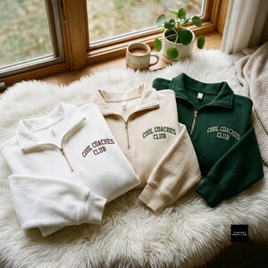 Könnte beinhalten: Drei Sweatshirts mit kurzem Reißverschluss in Weiß, Beige und Dunkelgrün. Jedes Kleidungsstück hat einen hohen Kragen mit goldenem Reißverschluss und den gestickten Text COOL COACHES CLUB in einer dunklen, geschwungenen Schrift.