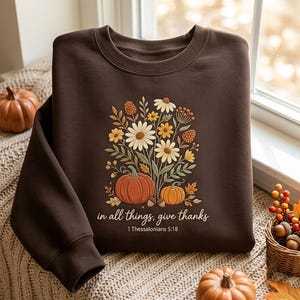 Puede incluir: Una sudadera de cuello redondo color marrón oscuro con un gráfico central de calabazas naranjas y un ramo de flores silvestres blancas, amarillas y naranjas. El texto dice "in all things, give thanks" seguido de "1 Thessalonians 5:18" en cursiva.