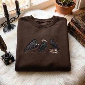 May include: A dark brown crewneck sweatshirt featuring an embroidered design of three black ravens. One raven holds an open book, another has a book balanced on its head, and the third stands atop a small stack of two books.