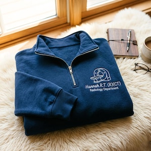 May include: A navy blue quarter-zip sweatshirt featuring a white embroidered graphic of a CT scanner. Below the graphic, the text reads "Hannah R.T. (R)(CT)" and "Radiology Department" in white lettering. The garment has a silver-toned zipper.