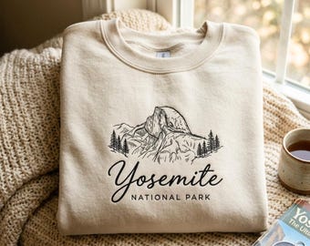 May include: A cream-colored crewneck sweatshirt featuring a black line-art illustration of a mountain peak with pine trees. Below the graphic, the text reads Yosemite NATIONAL PARK in a combination of script and sans-serif typography.