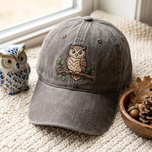 May include: A charcoal gray baseball cap featuring an embroidered design of a brown owl perched on a leafy branch. The cap has a structured crown with ventilation eyelets and a curved brim, showcasing detailed stitching throughout the fabric.