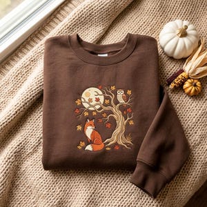 Könnte beinhalten: Ein dunkelbraunes Sweatshirt mit Rundhalsausschnitt und einer Stickerei, die einen Fuchs unter einem Baum mit einer darauf sitzenden Eule zeigt. Das Motiv enthält einen Vollmond und verstreute Herbstblätter in Orange-, Gelb- und Rottönen.
