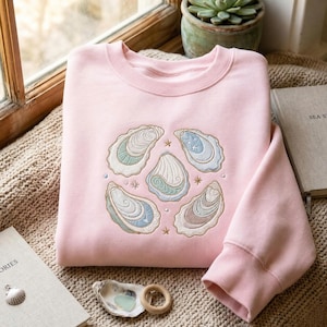May include: A light pink crewneck sweatshirt featuring an embroidered design of five oyster shells in shades of blue, green, and tan. The embroidery includes small star accents and dots arranged in a circular pattern on the center of the chest.