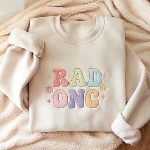 Op de afbeelding: Een crèmekleurige sweater met ronde hals en geborduurde tekst RAD ONC in een vrolijk, meerkleurig lettertype. Het ontwerp bevat kleine geborduurde bloem- en steraccenten in pasteltinten zoals roze, geel, blauw en paars.