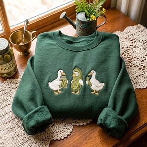 K&ouml;nnte beinhalten: Ein dunkelgr&uuml;nes Sweatshirt mit Rundhalsausschnitt und einer Stickerei, die zwei wei&szlig;e Enten und eine mittige Gew&uuml;rzgurkenfigur zeigt. Eine Ente h&auml;lt ein Glas Gew&uuml;rzgurken, w&auml;hrend die andere eine einzelne Gurke h&auml;lt. Stickerei in Wei&szlig;, Gr&uuml;n und Orange.