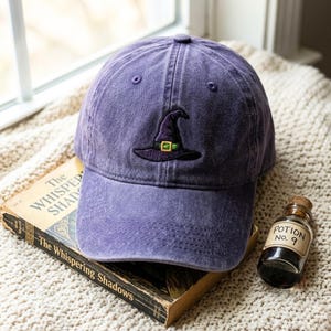 Può includere: Un berretto da baseball viola con finitura consumata che presenta un cappello da strega ricamato in viola scuro con una fascia verde e una fibbia dorata, posizionato su un libro intitolato The Whispering Shadows accanto a una boccetta Potion no. 9.