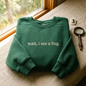 May include: A forest green crewneck sweatshirt featuring the embroidered text 'wait, i see a bug' in a light cream colour across the chest. The garment has long sleeves with ribbed cuffs and a ribbed neckline, presented in a simple, clean design.