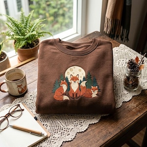 May include: A brown crewneck sweatshirt featuring an embroidered graphic of three orange foxes sitting in front of a large cream-coloured moon and dark green pine trees. The embroidery is centred on the chest of the garment.