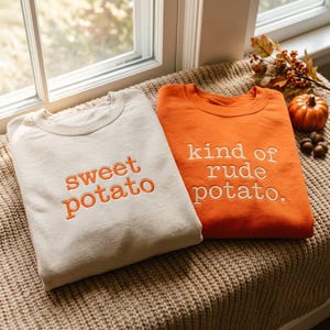 Może przedstawiać: Dwie bluzy z okrągłym dekoltem. Beżowa bluza posiada wyhaftowany pomarańczowy napis "sweet potato". Pomarańczowa bluza posiada wyhaftowany biały napis "kind of rude potato.". Oba ubrania mają długie rękawy i prążkowany dekolt.