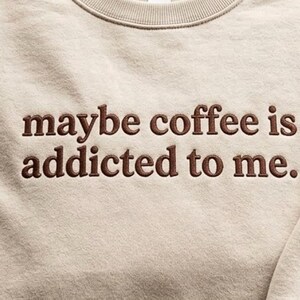 May include: A beige crewneck sweatshirt featuring dark brown embroidered text on the chest that reads, maybe coffee is addicted to me. The garment has a ribbed neckline and a soft, textured fabric finish across the front panel.