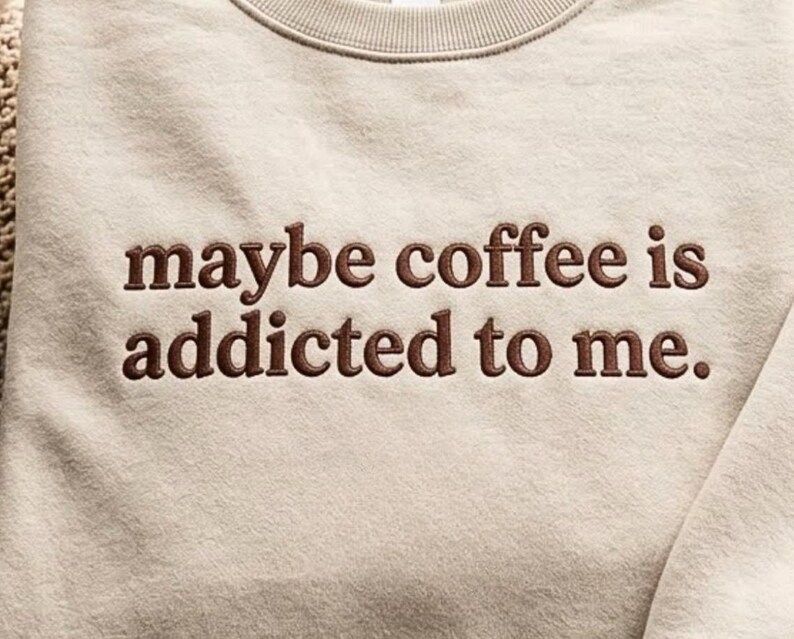 May include: A beige crewneck sweatshirt featuring dark brown embroidered text on the chest that reads, maybe coffee is addicted to me. The garment has a ribbed neckline and a soft, textured fabric finish across the front panel.