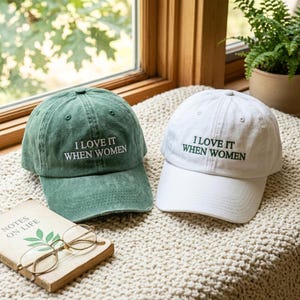 Peut inclure: Deux casquettes de baseball, l'une en vert délavé et l'autre en blanc, arborant toutes deux le texte brodé "I LOVE IT WHEN WOMEN" dans une police sans empattement centrée. Chaque casquette possède une visière courbée et des œillets.