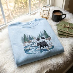Peut inclure: Un sweat-shirt bleu clair à col rond orné d'une broderie représentant une scène naturelle. Le motif montre une rivière bleue sinueuse, des rochers gris, des pins verts et trois ours noirs marchant dans un paysage enneigé aux détails blancs et bruns.