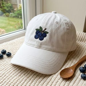May include: A white baseball cap featuring an embroidered design of a cluster of blue blueberries with green leaves on the front panel. The cap has a curved brim with visible stitching and a button at the top centre of the crown.