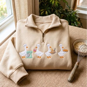 May include: A beige quarter-zip sweatshirt featuring an embroidered design of four white geese wearing chef hats. The geese are depicted baking, with one using a mint green stand mixer, one holding a cupcake, one carrying bread, and one holding a pie.