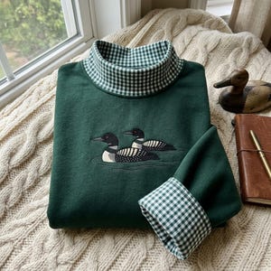 Puede incluir: Una sudadera de manga larga de color verde oscuro con un bordado de dos colimbos nadando en el agua. La prenda tiene un cuello alto y puños en las mangas forrados con una tela de patrón de cuadros vichy en blanco y verde oscuro.