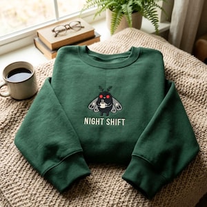 May include: A dark green crewneck sweatshirt featuring an embroidered graphic of a black moth with red eyes holding a steaming white mug. Below the moth, the text NIGHT SHIFT is embroidered in white. The sweatshirt has long sleeves and ribbed cuffs.