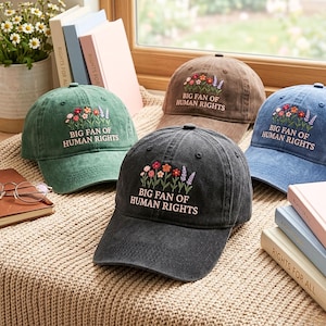 May include: Four baseball caps in green, brown, blue, and black, each featuring embroidered colorful flowers and the text "BIG FAN OF HUMAN RIGHTS" on the front. The caps are arranged with books and a pair of glasses on a textured surface.