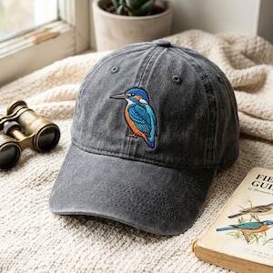 May include: A charcoal grey baseball cap featuring an embroidered kingfisher bird on the front panel. The bird has a bright blue head and wings with an orange chest and a long, pointed black beak. The cap has a curved brim and stitched eyelets.