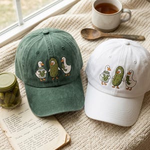 May include: Two baseball caps, one in dark green and one in white, each featuring an embroidered design of two white ducks and a central green pickle character. The left duck holds a jar of pickles, while the right duck holds a single pickle.