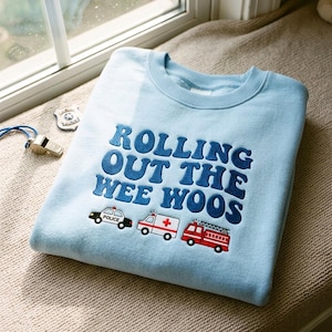 Könnte beinhalten: Ein hellblaues Sweatshirt mit Rundhalsausschnitt und dunkelblau gesticktem Text ROLLING OUT THE WEE WOOS. Unter dem Text befinden sich kleine Stickereien eines schwarz-weißen Polizeiautos, eines weißen Krankenwagens mit rotem Kreuz und eines roten Feuerwehrautos.