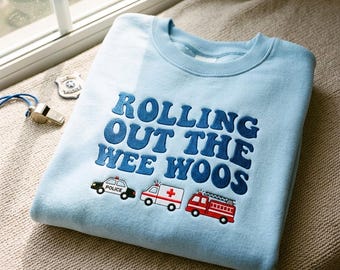 May include: A light blue crewneck sweatshirt featuring dark blue embroidered text that reads ROLLING OUT THE WEE WOOS. Below the text are small embroidered graphics of a black and white police car, a white ambulance with a red cross, and a red fire truck.